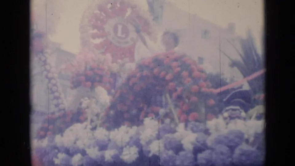 The 1962 Cannes footage lingers on a dense floral float, with red blossoms, pale flowers, and a Lions Club emblem visible above.