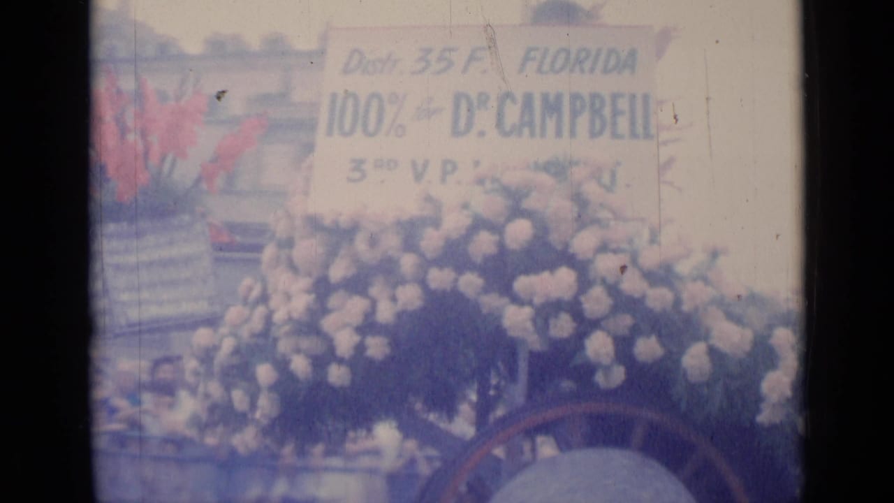 Parade float with flowers, a Florida Dr. Campbell sign, and people partly visible behind it.
