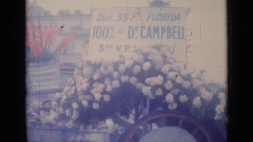 Cannes 1962 parade float frame with dense flowers, a readable Florida Dr. Campbell sign, and onlookers or riders partly obscured.