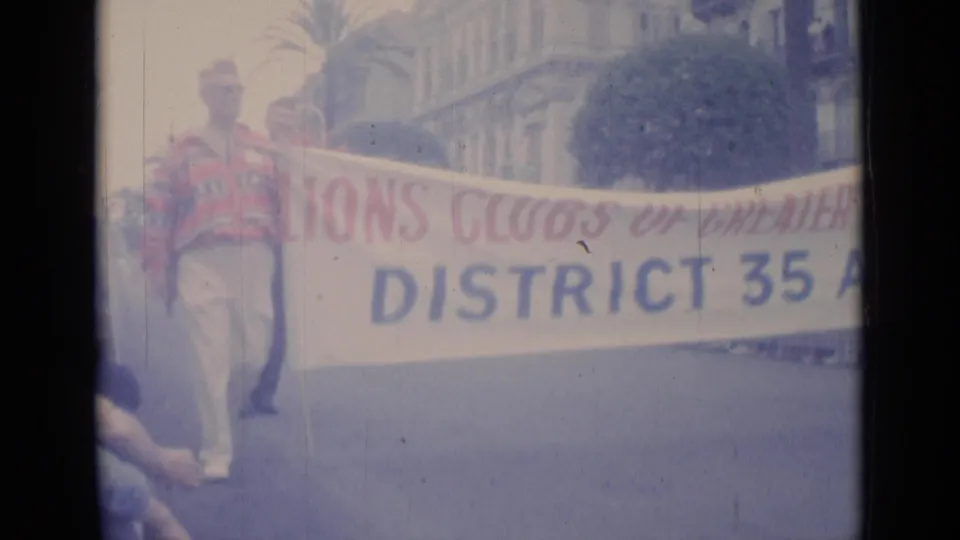 1962 Cannes parade frame with men carrying a Lions Clubs of Cannes District 35A banner in a public street procession.