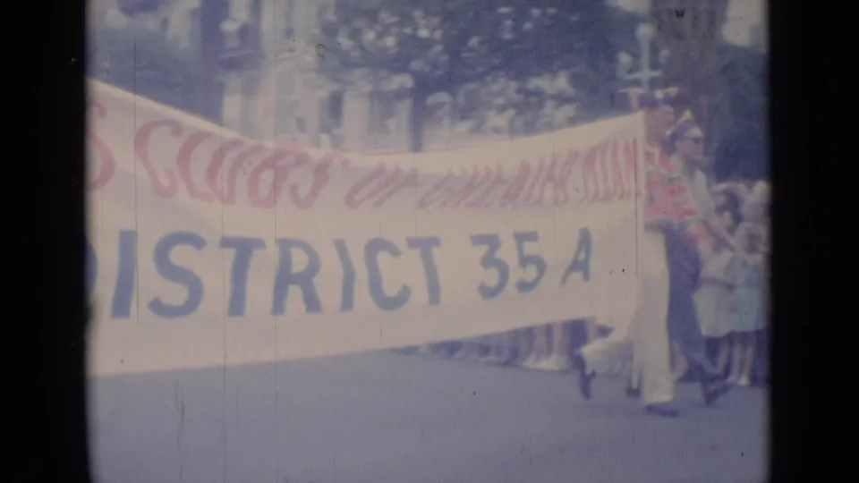 Cannes home-movie frame showing the District 35A parade banner stretched across the street with walkers and bystanders nearby.