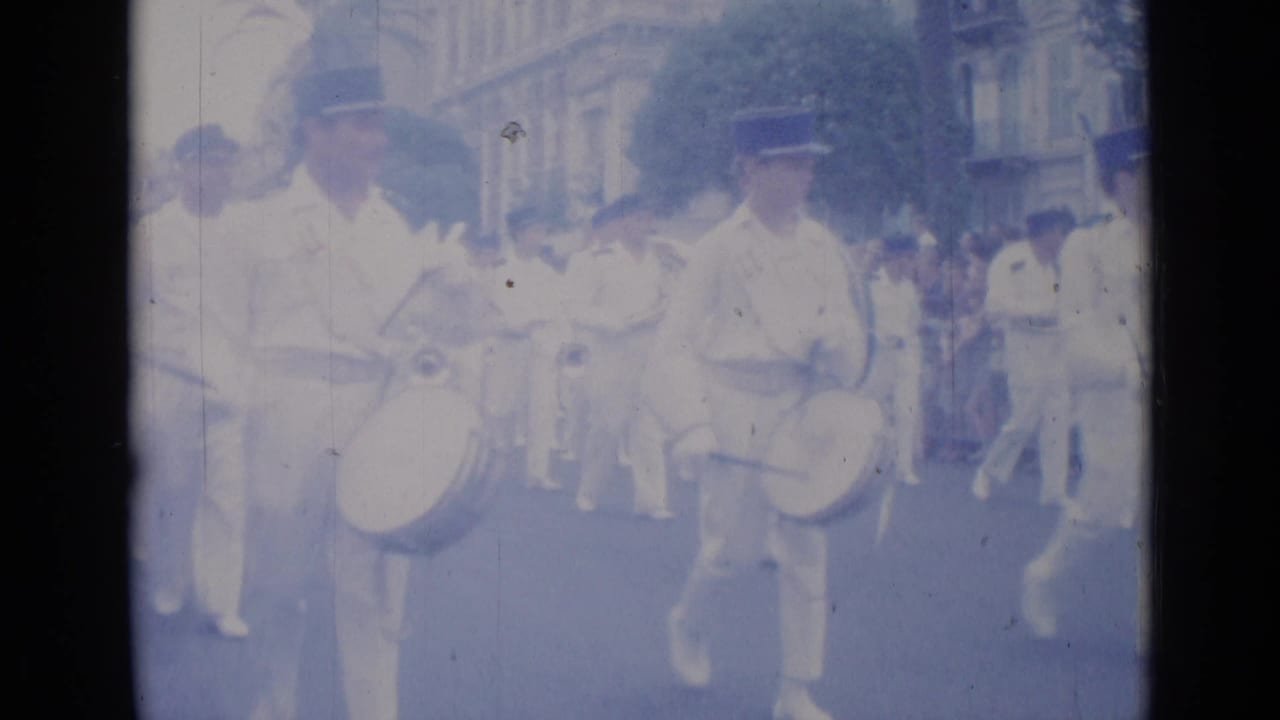 White-uniformed marching band members with drums move down a city street.