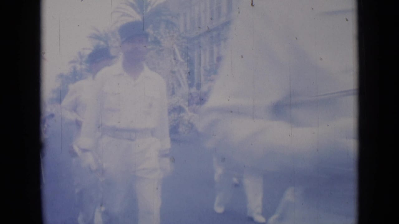 Cannes 1962 home-movie frame showing a man in a white uniform and cap walking beside a light-colored car on a palm-lined street.