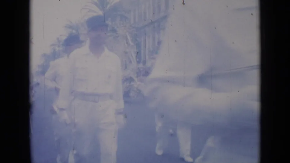 Cannes 1962 home-movie frame showing a man in a white uniform and cap walking beside a light-colored car on a palm-lined street.