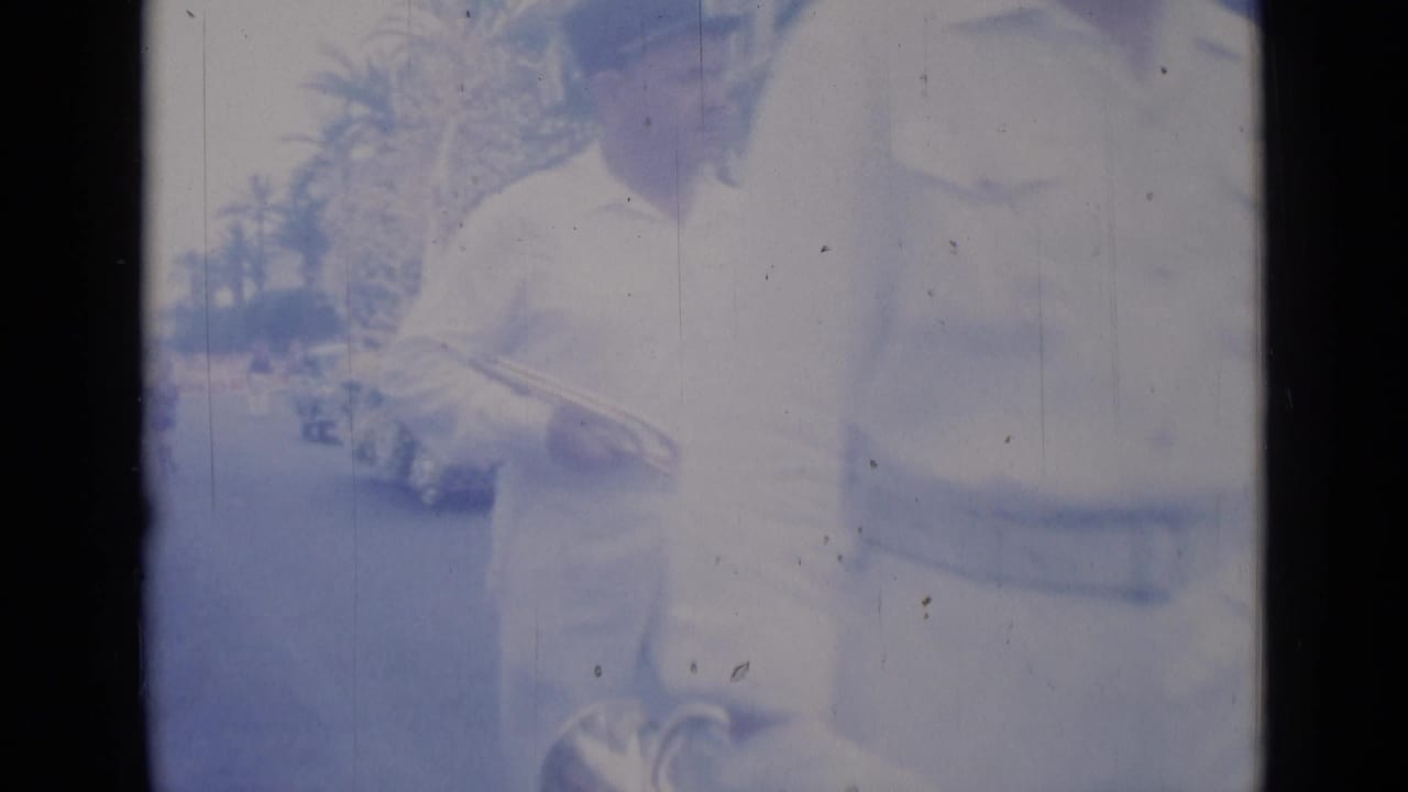 Uniformed man stands or marches near a bright car with palm trees in the background.