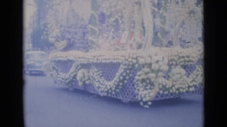 Cannes 1962 frame showing a large flower-covered float with figures on top, moving along a palm-lined city street.