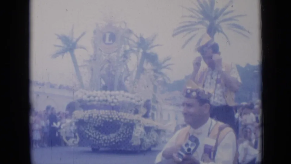 1962 Cannes parade scene with a man in ceremonial vest and hat holding a camera, with floral displays and palm trees behind him.