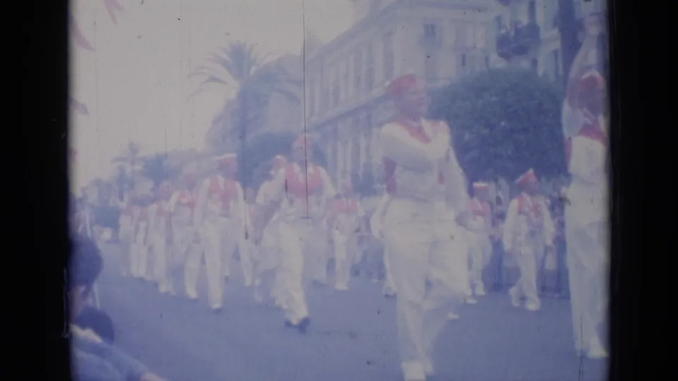 A Cannes parade group in white clothing and red sashes advances past roadside spectators and mid-century buildings.