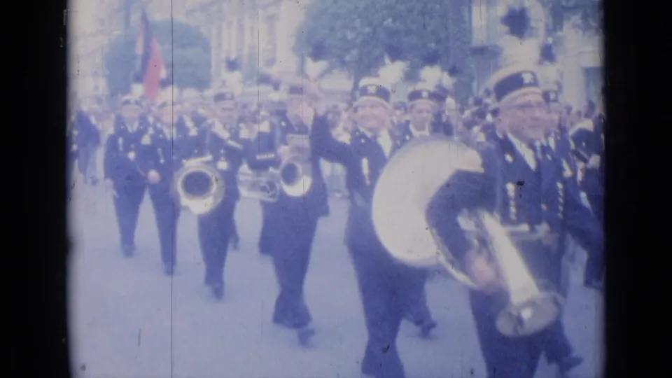 Uniformed musicians, including a large drum player and brass players, march through Cannes as the home-movie camera watches from street level.