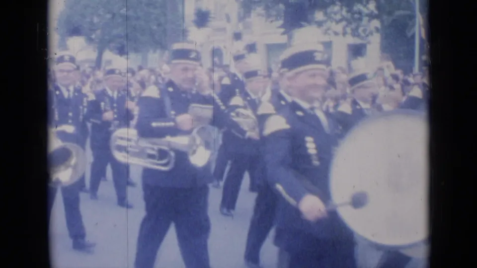 A smiling uniformed drummer passes close to the camera while other band members with brass instruments continue through Cannes.