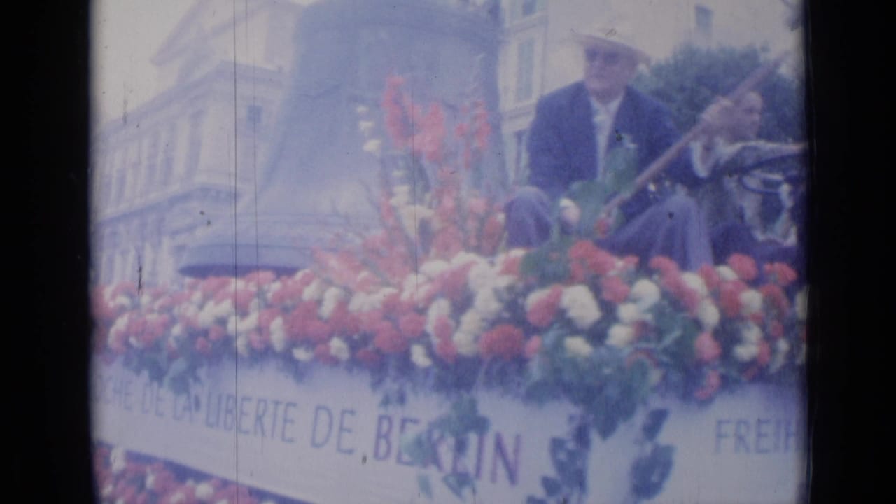 1962 Cannes parade frame with a suited man on a red and white floral float bearing Berlin freedom signage.