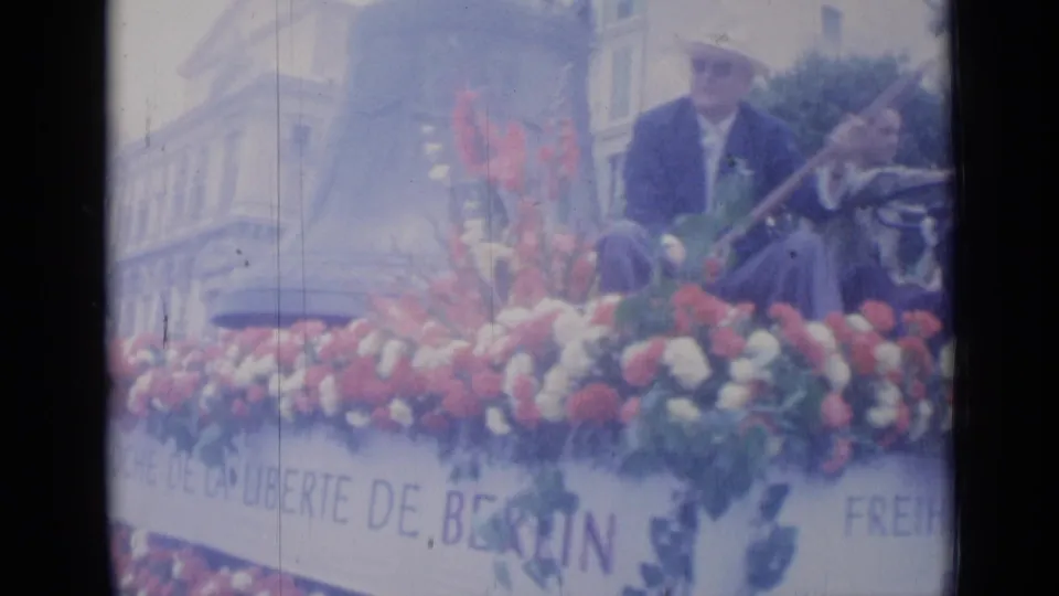 1962 Cannes parade frame with a suited man on a red and white floral float bearing Berlin freedom signage.