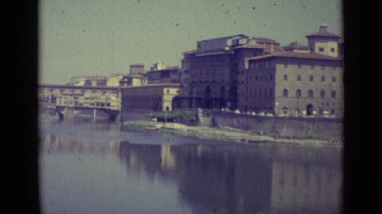 1962 8mm view of Florence buildings beside the Arno River, with a bridge at left and soft reflections in the water.