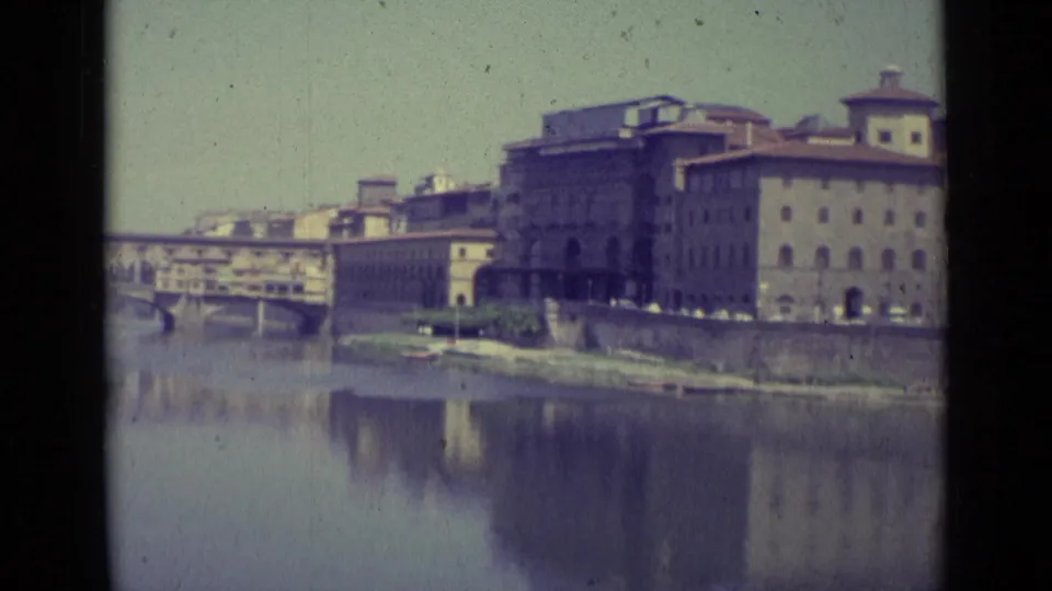 1962 8mm view of Florence buildings beside the Arno River, with a bridge at left and soft reflections in the water.
