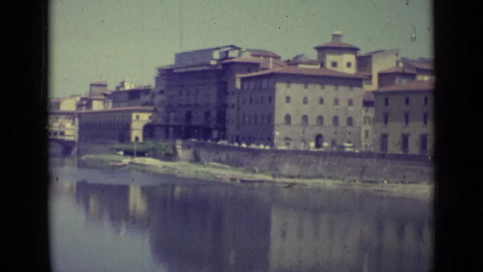 Florence riverfront buildings in 1962 home-movie footage, with pale sky, rooflines, and reflections across the Arno River.