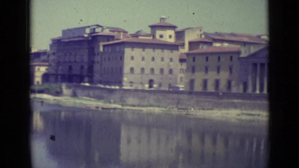 1962 Florence waterfront view with tan buildings, river reflections, and part of a classical columned facade entering at right.