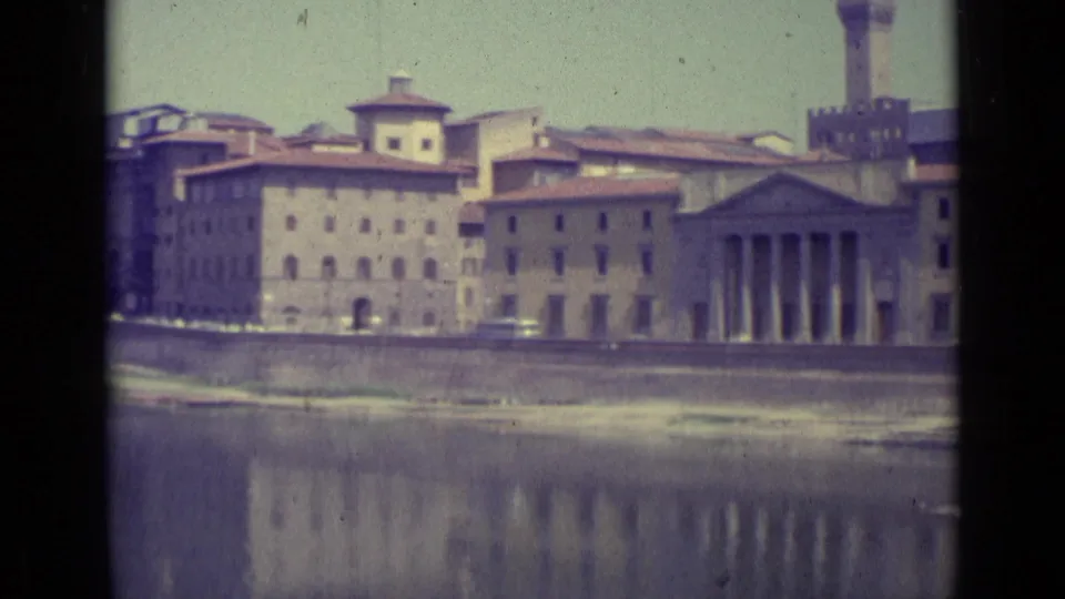 Florence in 1962 with a classical columned facade, adjoining tan buildings, and their reflections across the Arno River.