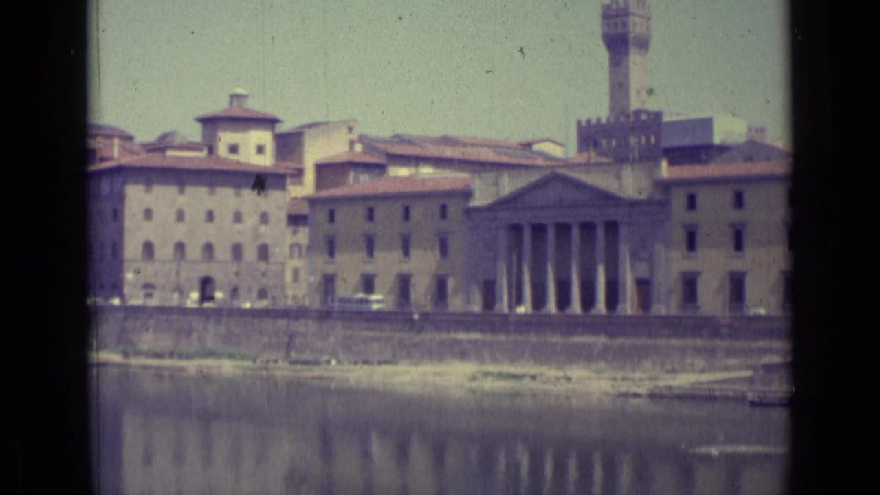 1962 Florence view across the river toward historic buildings, a columned facade, and a tall tower, framed by 8mm film edges.