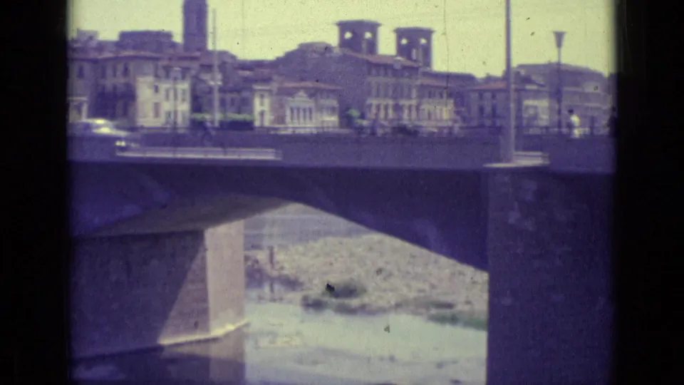 1962 Florence bridge scene with cars and small pedestrian figures moving past buildings above a shallow river channel.