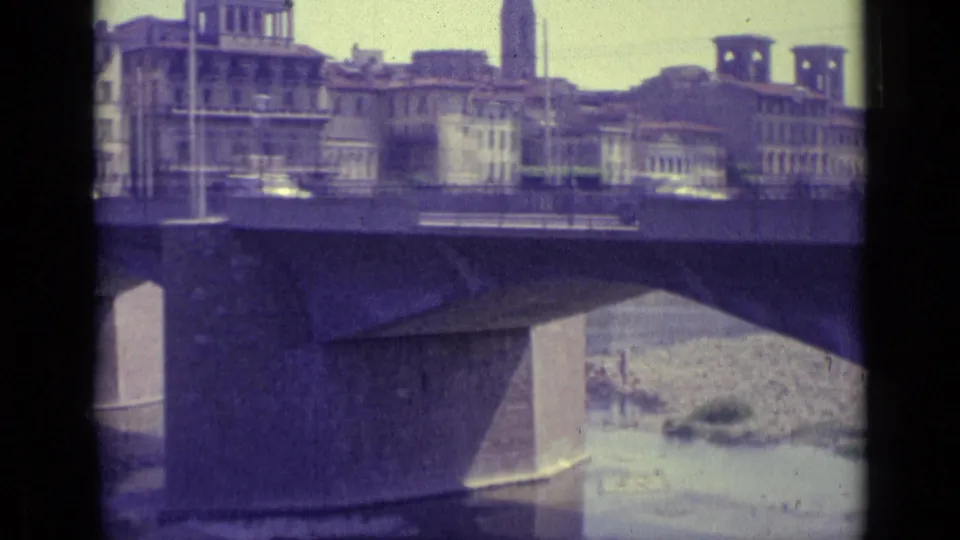 Florence bridge traffic in 1962, with cars crossing above the river and historic city buildings visible beyond the railing.