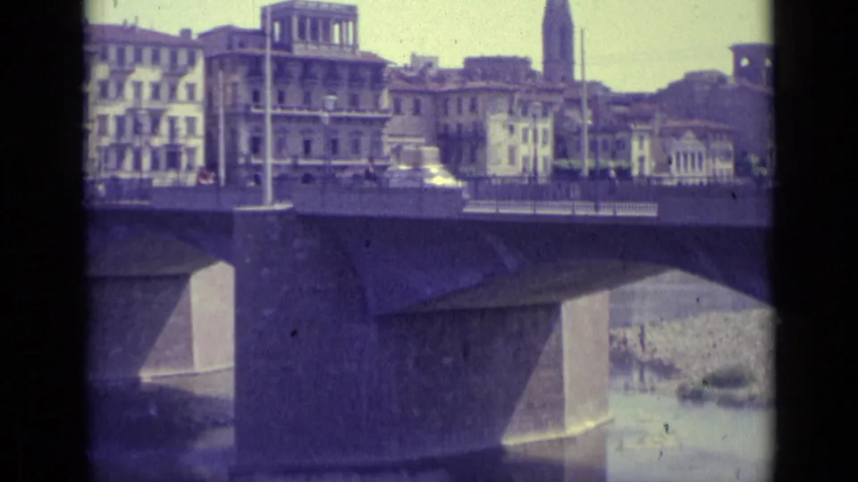 1962 Florence 8mm frame showing cars on a bridge above river water, with multi-story riverside buildings in the background.