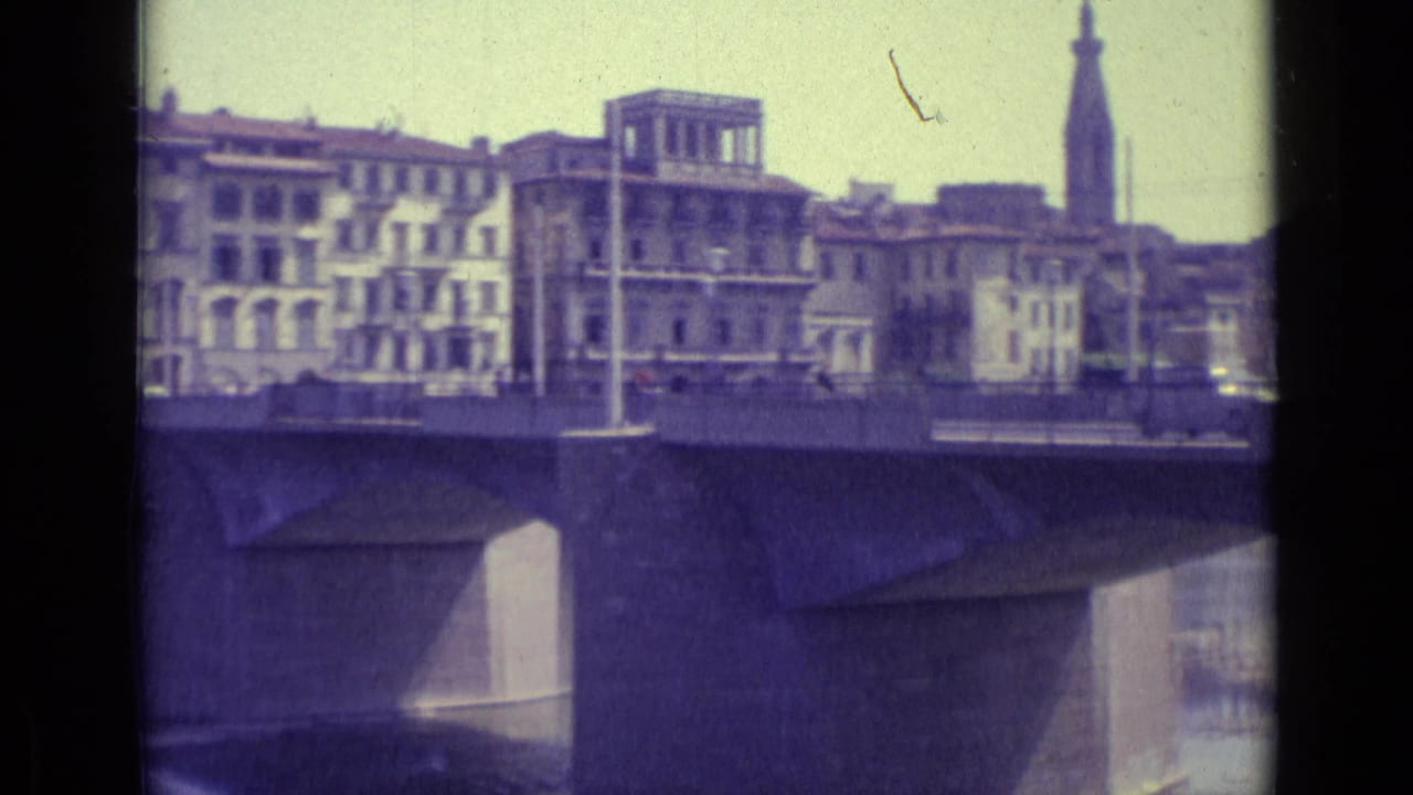 Bridge traffic passes in front of riverfront buildings and a tall tower.