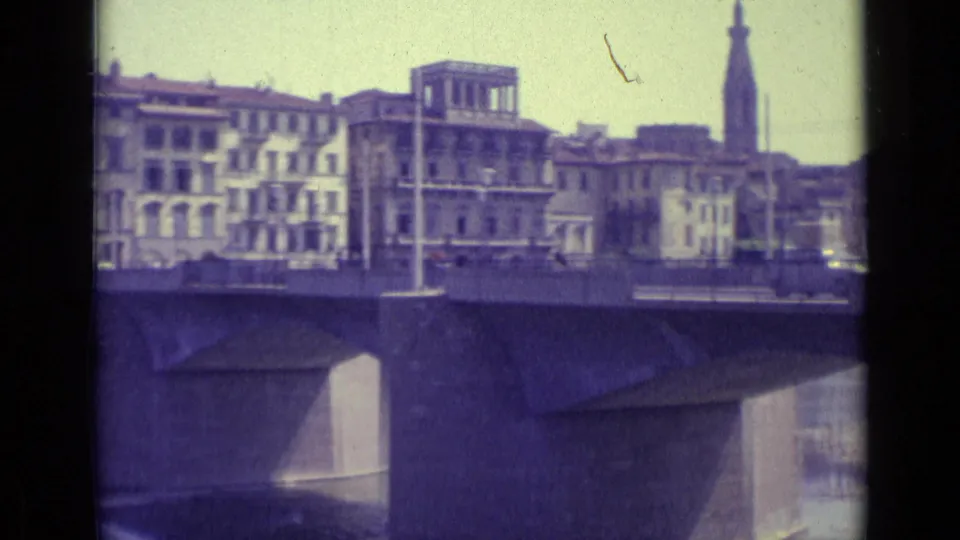 1962 Florence view of a bridge over the river, with cars on the deck and a tall tower visible beyond the waterfront buildings.