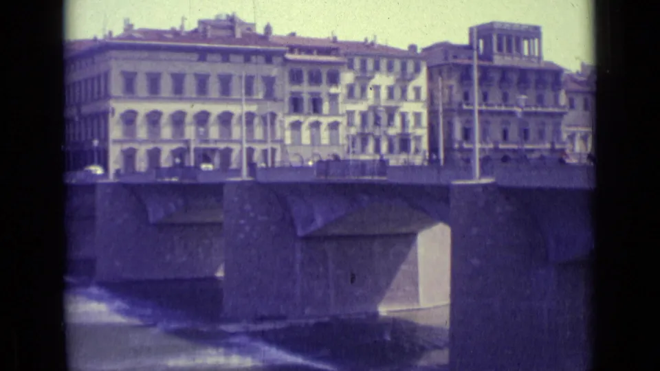 1962 Florence 8mm frame with cars crossing the bridge, arched building facades, lampposts, and river water below.