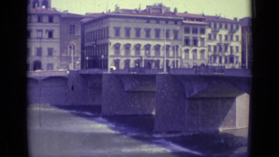 1962 Florence riverfront frame showing bridge piers, moving cars on the bridge, and closely spaced buildings along the far bank.