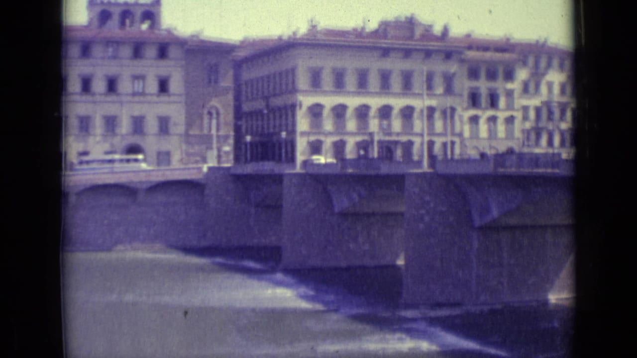 1962 Florence riverfront view with windowed buildings, a stone bridge, water, and faint vehicles in faded 8mm color.