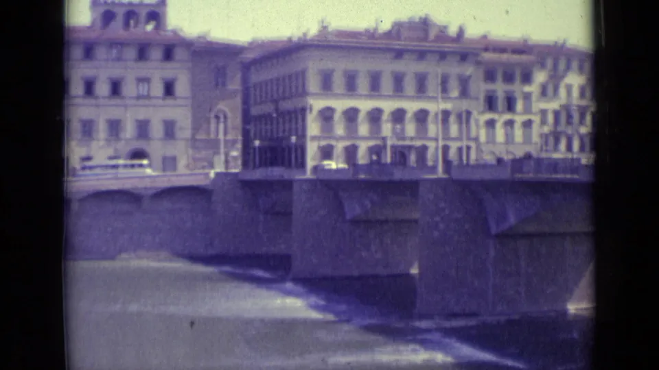 1962 Florence riverfront view with windowed buildings, a stone bridge, water, and faint vehicles in faded 8mm color.