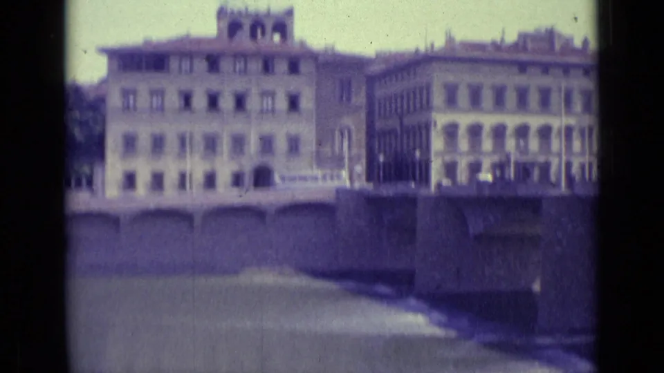 Florence riverbank buildings and a stone bridge appear across the water in a blurred 1962 8mm travel frame.