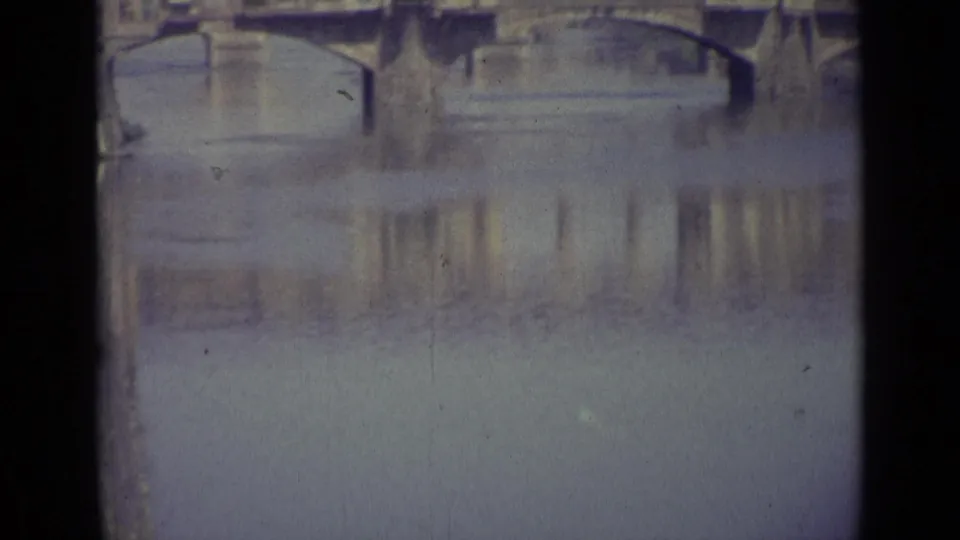 1962 Florence home-movie frame showing a bridge with buildings along the span reflected in calm water.