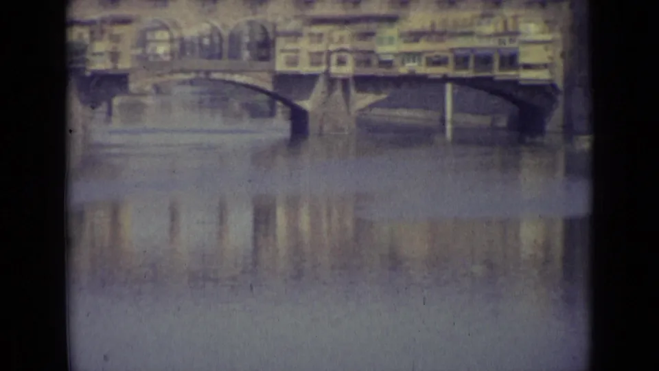 A soft 1962 8mm view of a Florence bridge, with buildings over the span and reflections below.