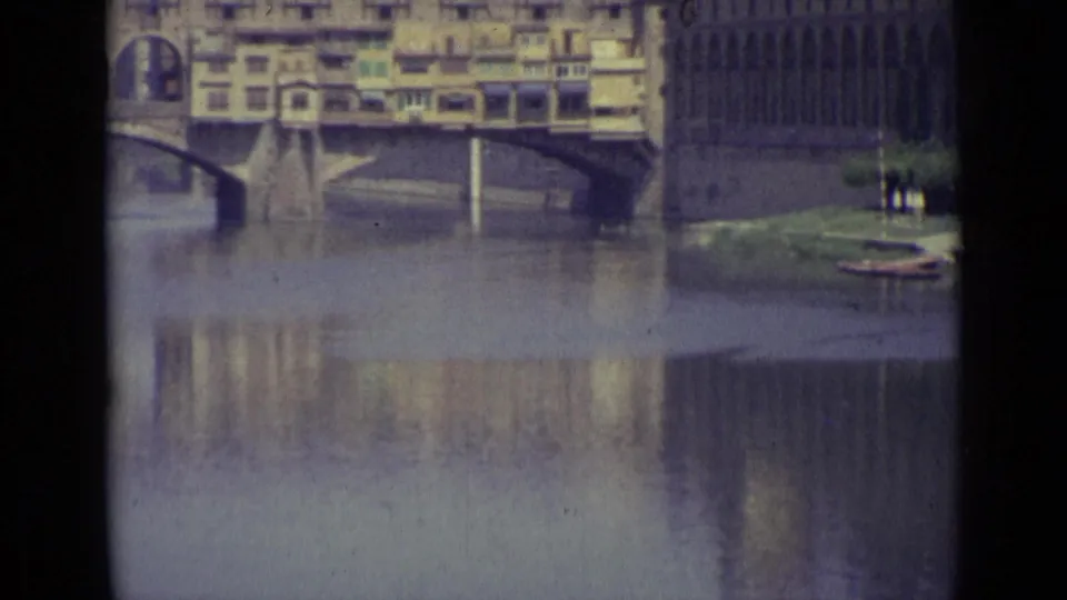 Final 1962 Florence frame with the bridge, reflected buildings, a riverside facade, and a small grassy edge.
