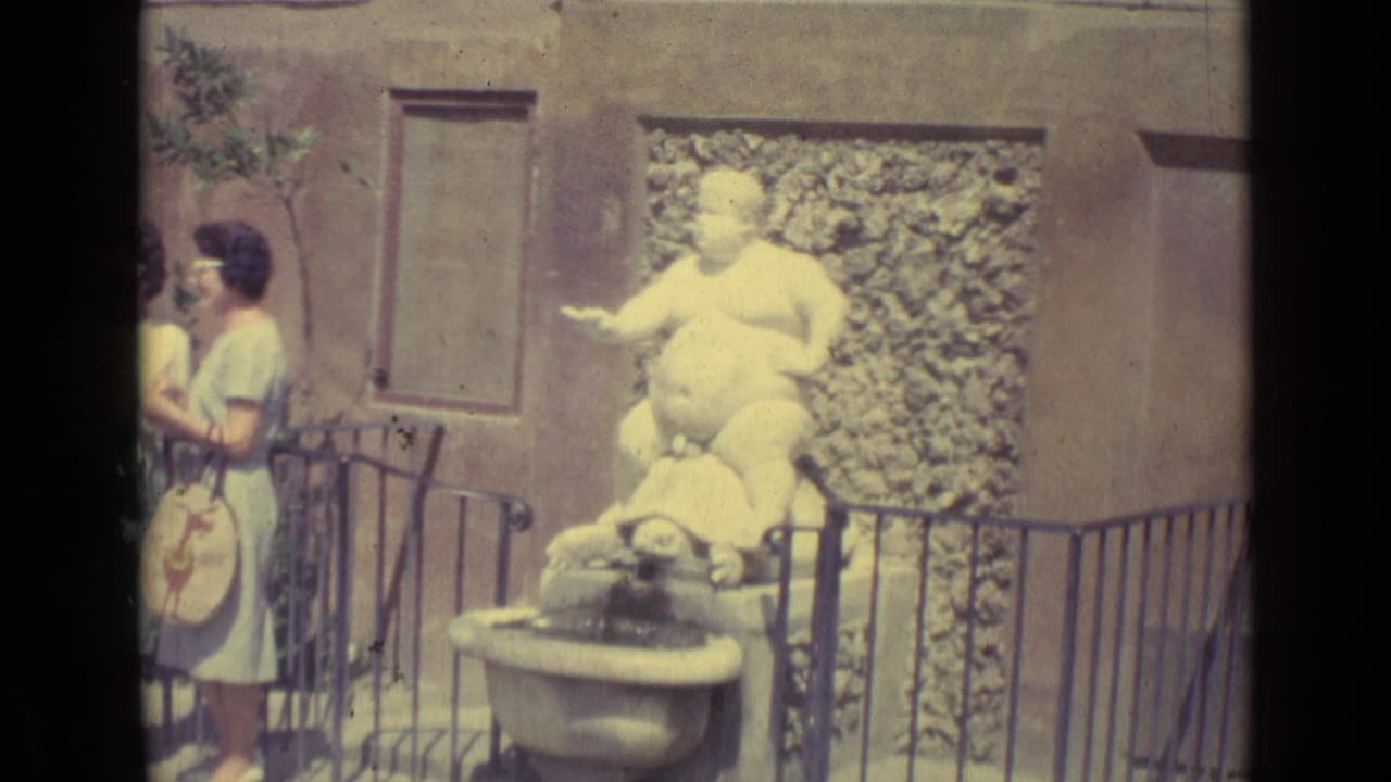 1962 Florence home-movie frame showing a bright white seated figure on a turtle fountain near a black railing, with one woman partly at left.