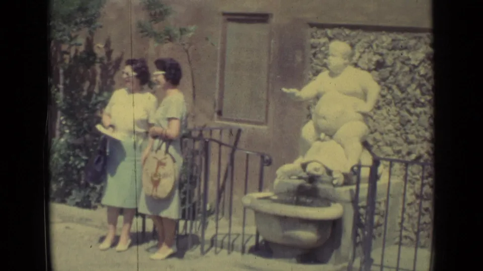 Two adult women in summer dresses stand beside a railing near the white turtle fountain sculpture in Florence, filmed on faded 8mm color stock.