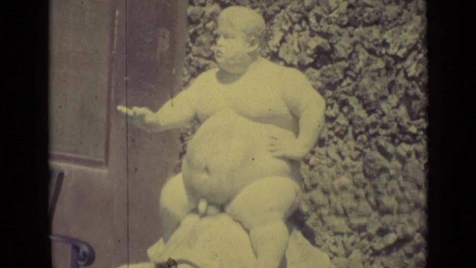 1962 Florence 8mm frame showing a plump nude boy statue riding a turtle near a textured stone wall and metal railing.