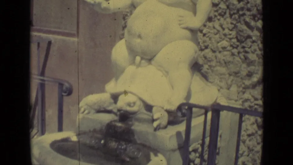1962 Florence home-movie frame looking down across the boy-on-turtle sculpture near railings and a fountain basin.