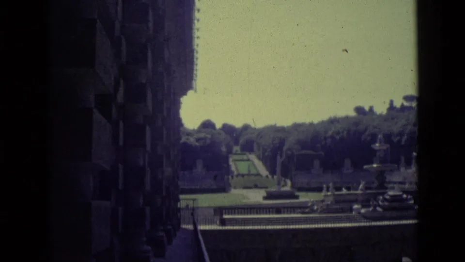 Wider 1962 Florence 8mm view of a formal garden area with fountain, path, trees, railing, and building edge.