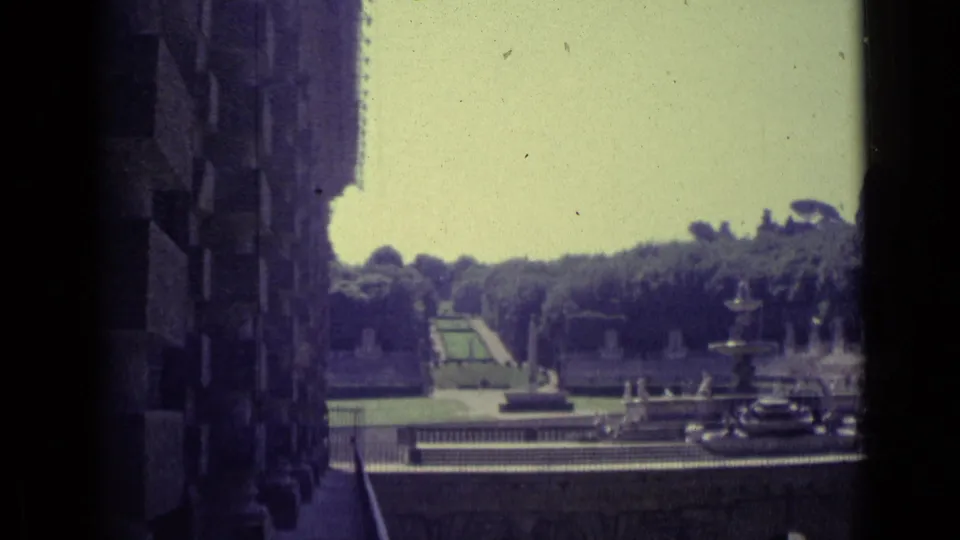1962 Florence 8mm frame showing a formal garden, fountain, balustrade, and distant trees from beside a dark stone facade.