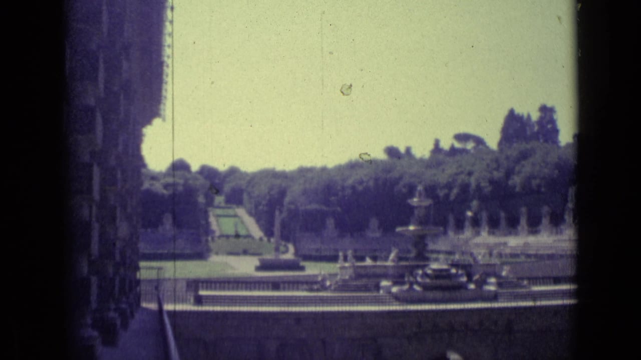 Garden terrace with fountains, trees, and dark cars near the foreground road.