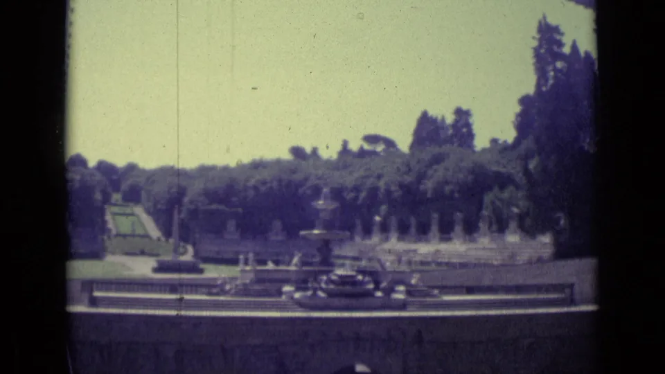 1962 Florence 8mm frame looking across a formal garden, fountain spray, tree line, and foreground roadway with dark cars.