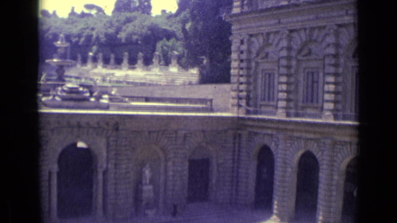 Rusticated stone building with arched colonnade, shuttered windows, fountain, and trees.