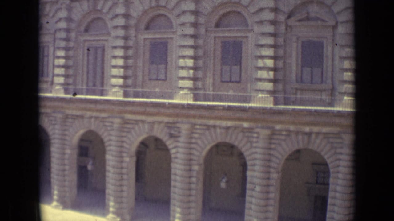 Stone arcade and shuttered windows viewed through vertical frame bars.