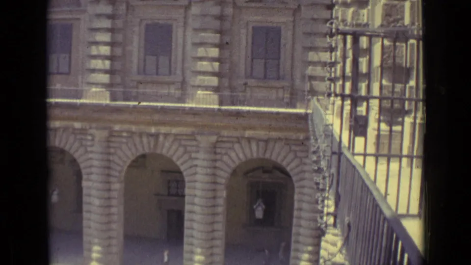 1962 Florence home-movie frame showing a sunlit historic facade with arched openings, tall windows, and a dark iron railing.