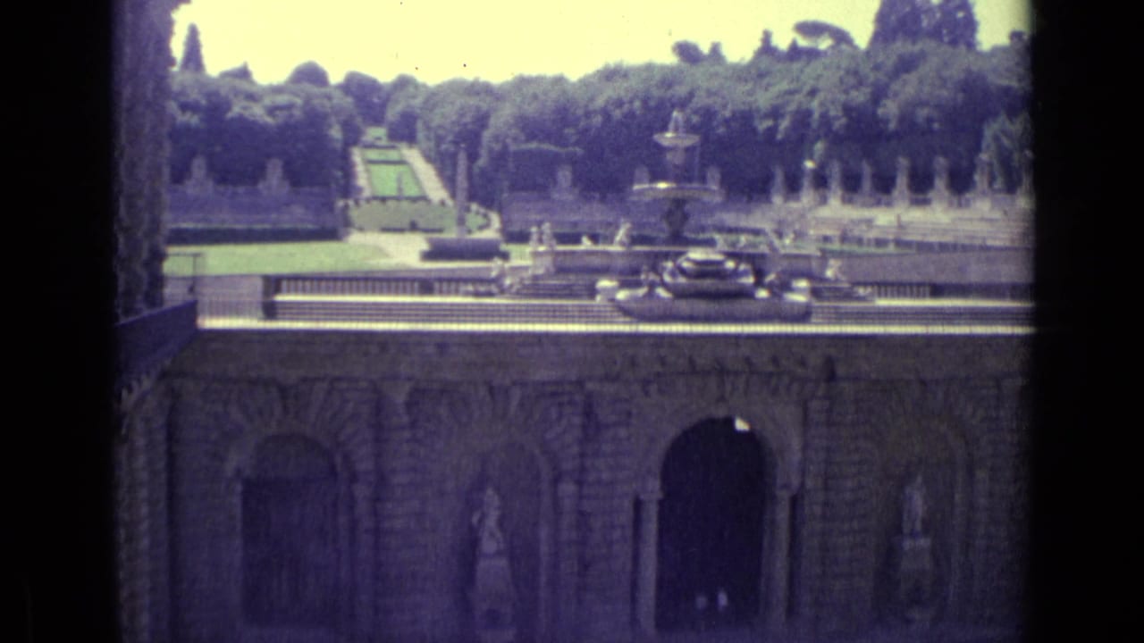 Formal garden with terraces, fountain, statues, trees, and cars beyond a stone wall.