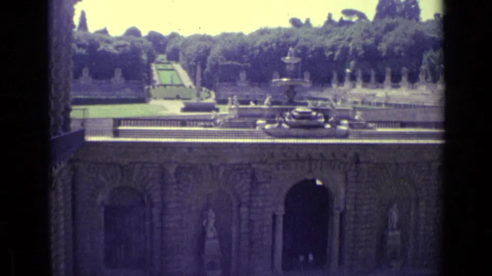 1962 Florence frame looking across a historic wall toward a formal garden with fountain spray, statues, terraces, trees, and parked cars.