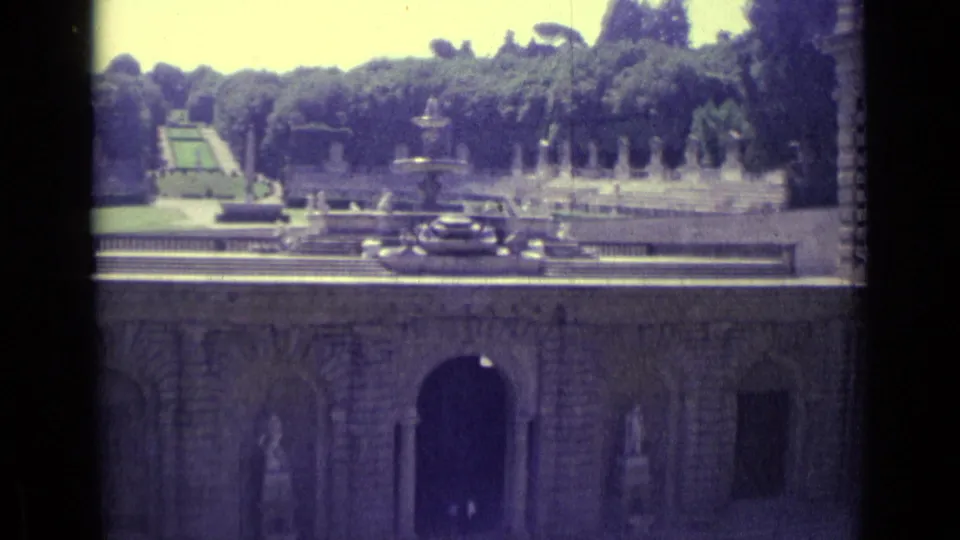 1962 Florence home-movie view of a formal garden and fountain above arched masonry, with statues, trees, and parked cars visible.