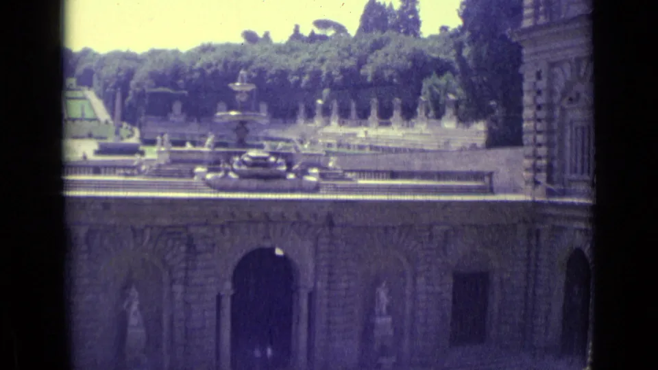 1962 Florence frame showing a formal garden with fountain spray and trees above a historic arched facade and the edge of a masonry building.